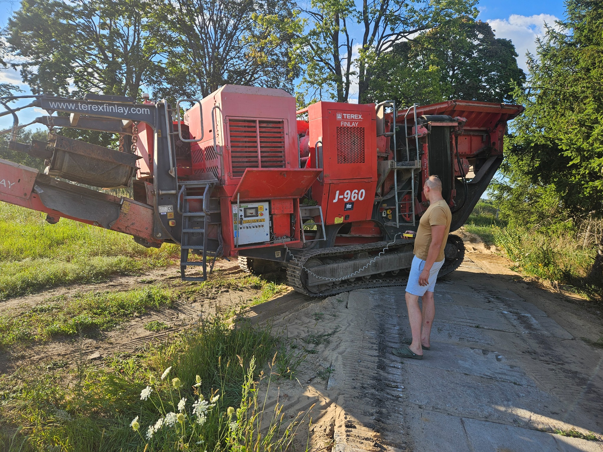 Czerwona maszyna do kruszenia kamieni Terex Finlay J-960 na gąsienicach, z mężczyzną w tle, na terenie zielonym z drzewami i śladami opon.