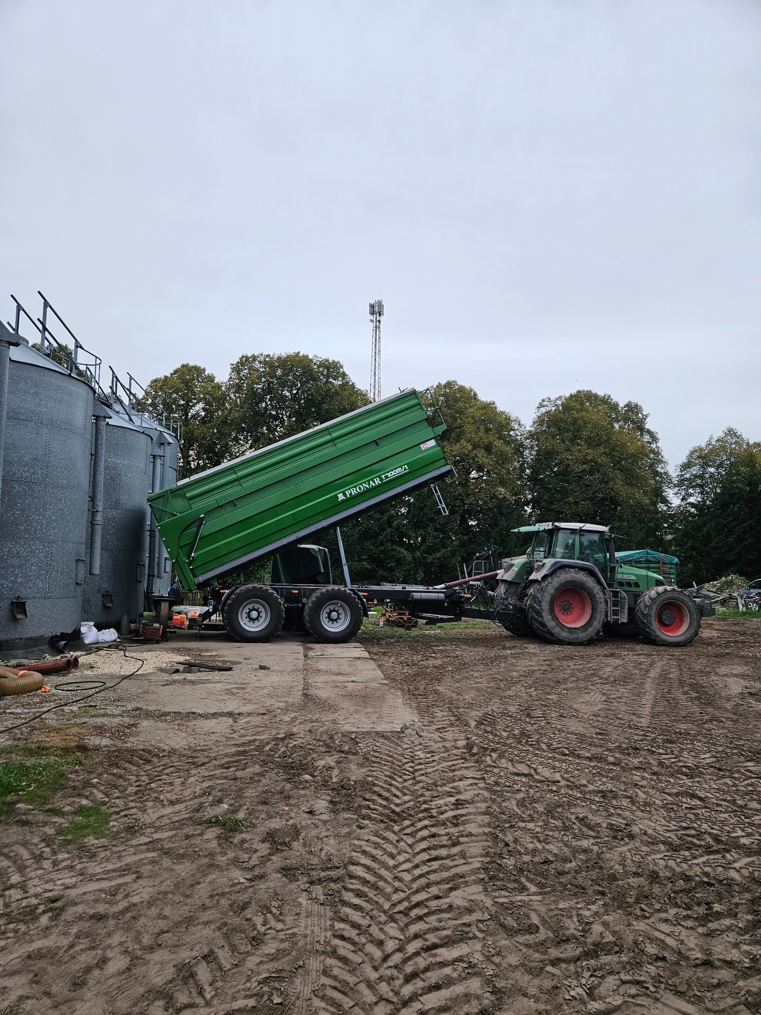Zielony traktor Fendt z podniesioną przyczepą Pronar T700M/1, obok silosów zbożowych na ubitym gruncie. Widoczne ślady opon i drzewa w tle.