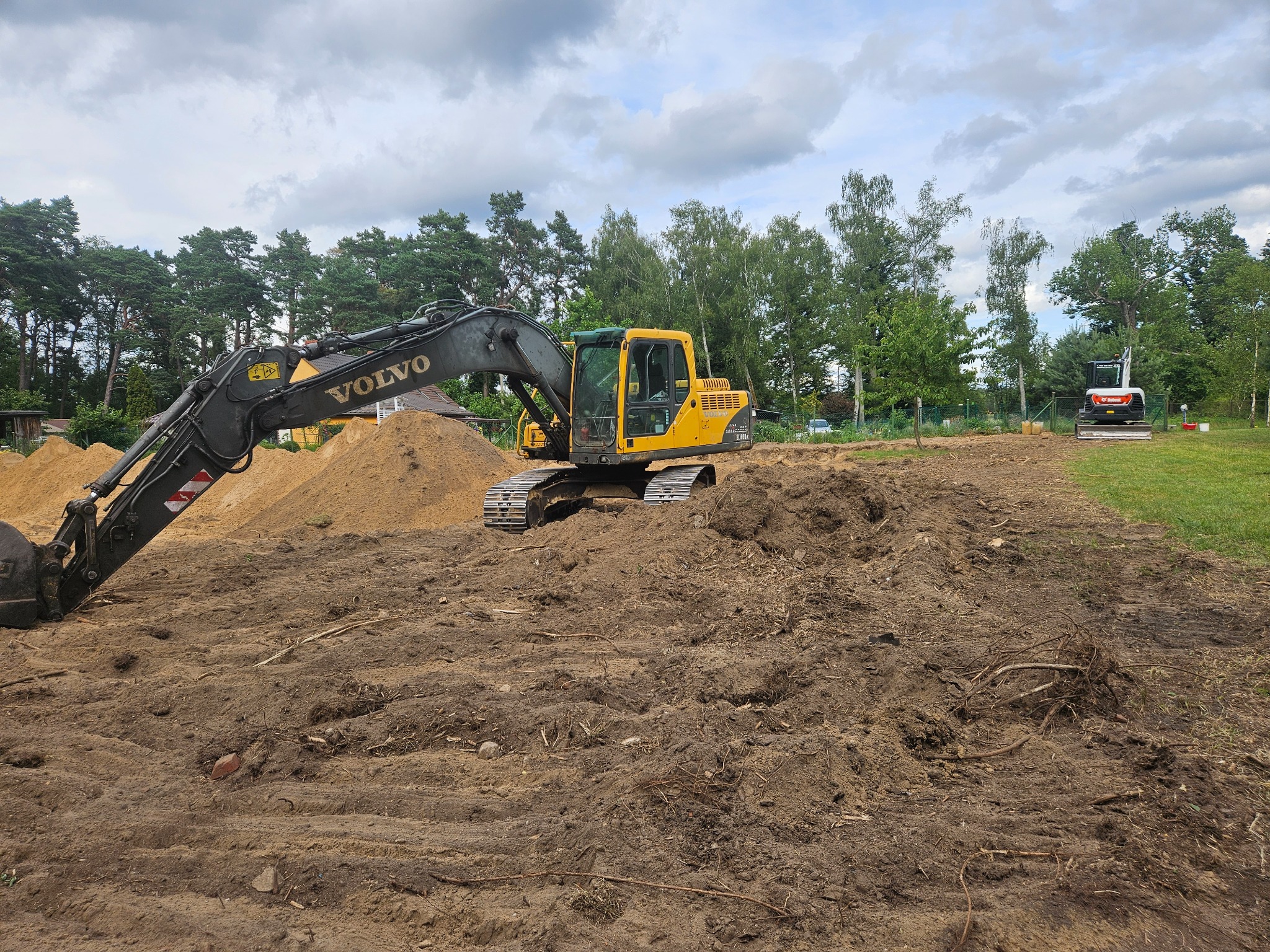 Żółta koparka Volvo na placu budowy, w tle drzewa i niebo z chmurami. Widoczny drugi, mniejszy sprzęt. Ślady pracy ziemnej, świeżo przekopana ziemia.