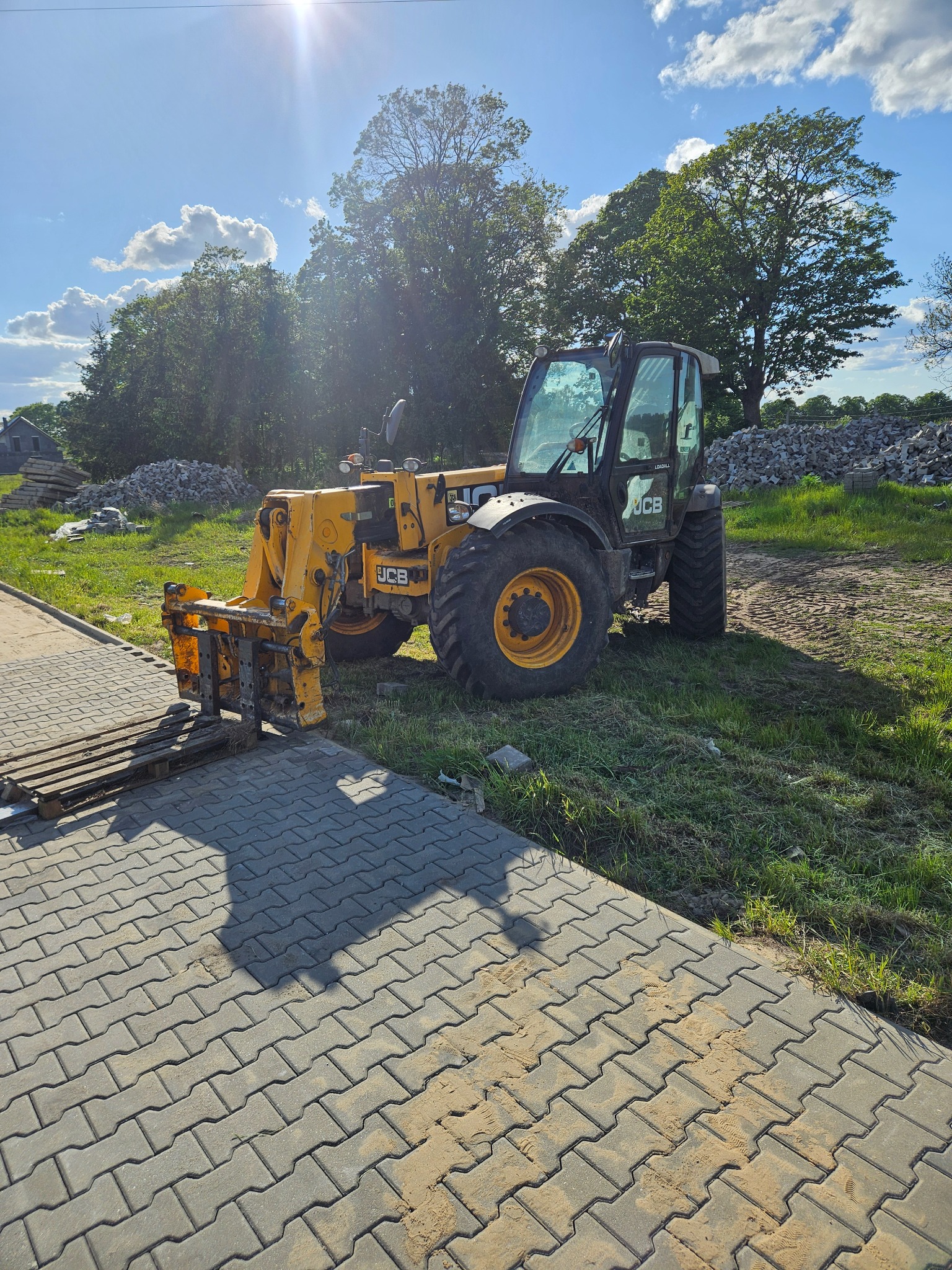 Żółta ładowarka teleskopowa JCB na placu budowy, z paletą kostki brukowej na widłach, w tle drzewa i sterta gruzu. Słoneczny dzień.