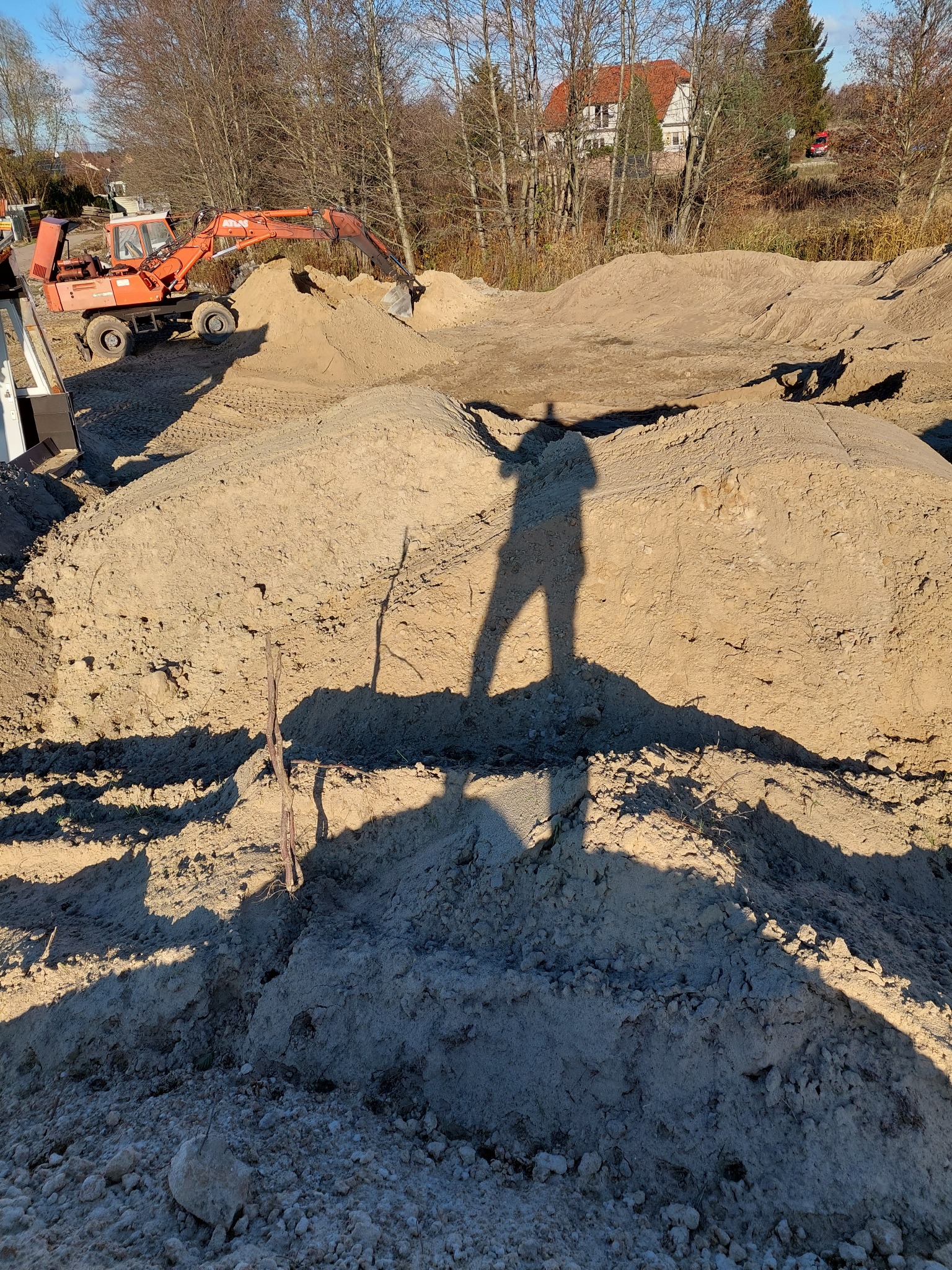 Pomarańczowa koparka obok hałdy piasku, cień osoby na piasku, w tle drzewa i budynek z czerwoną dachówką.