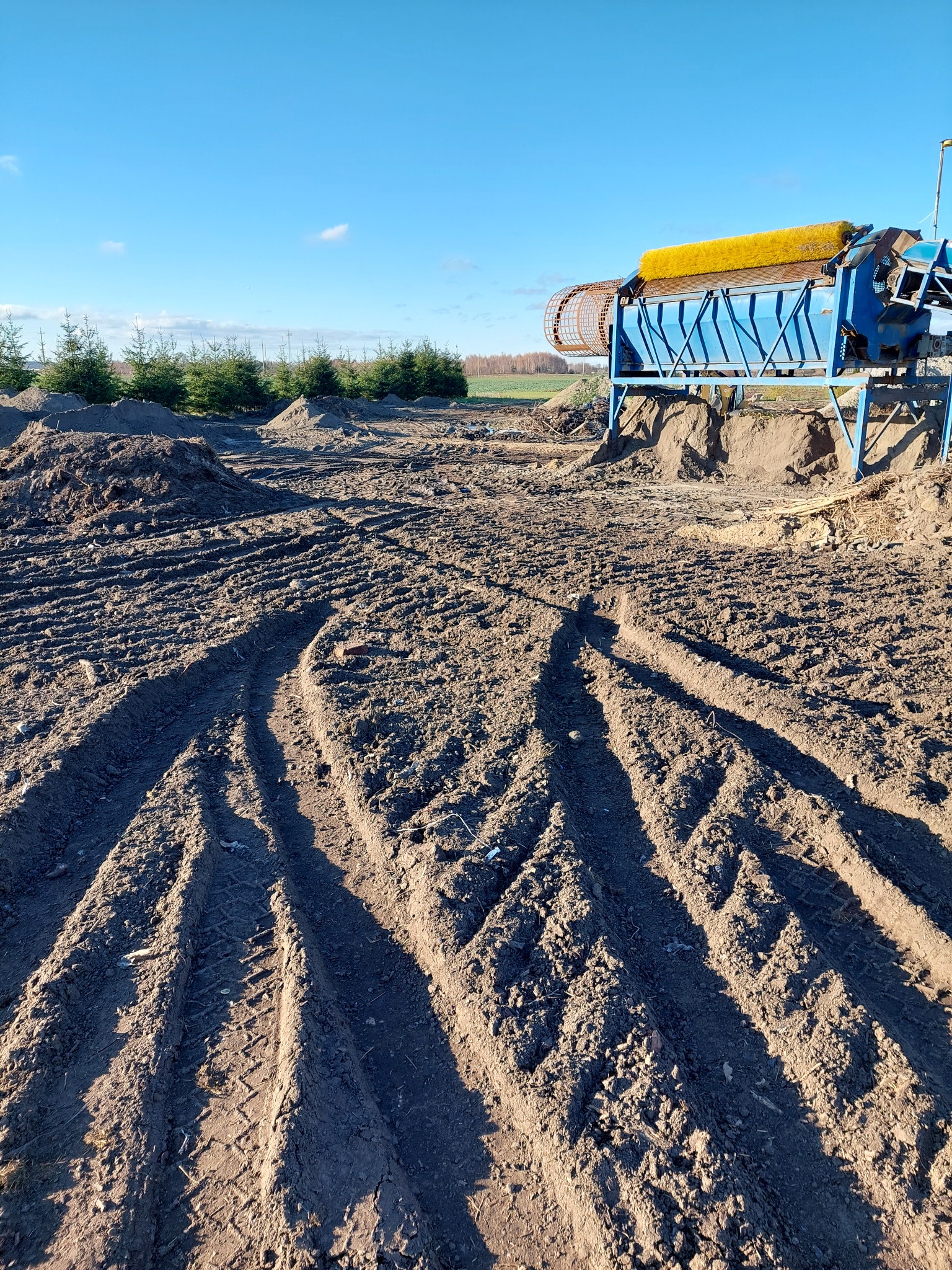 Nierówny teren po robotach ziemnych z widocznymi śladami opon i niebieską przesiewarką do ziemi z żółtą szczotką na tle małych drzew iglastych i błękitnego nieba.
