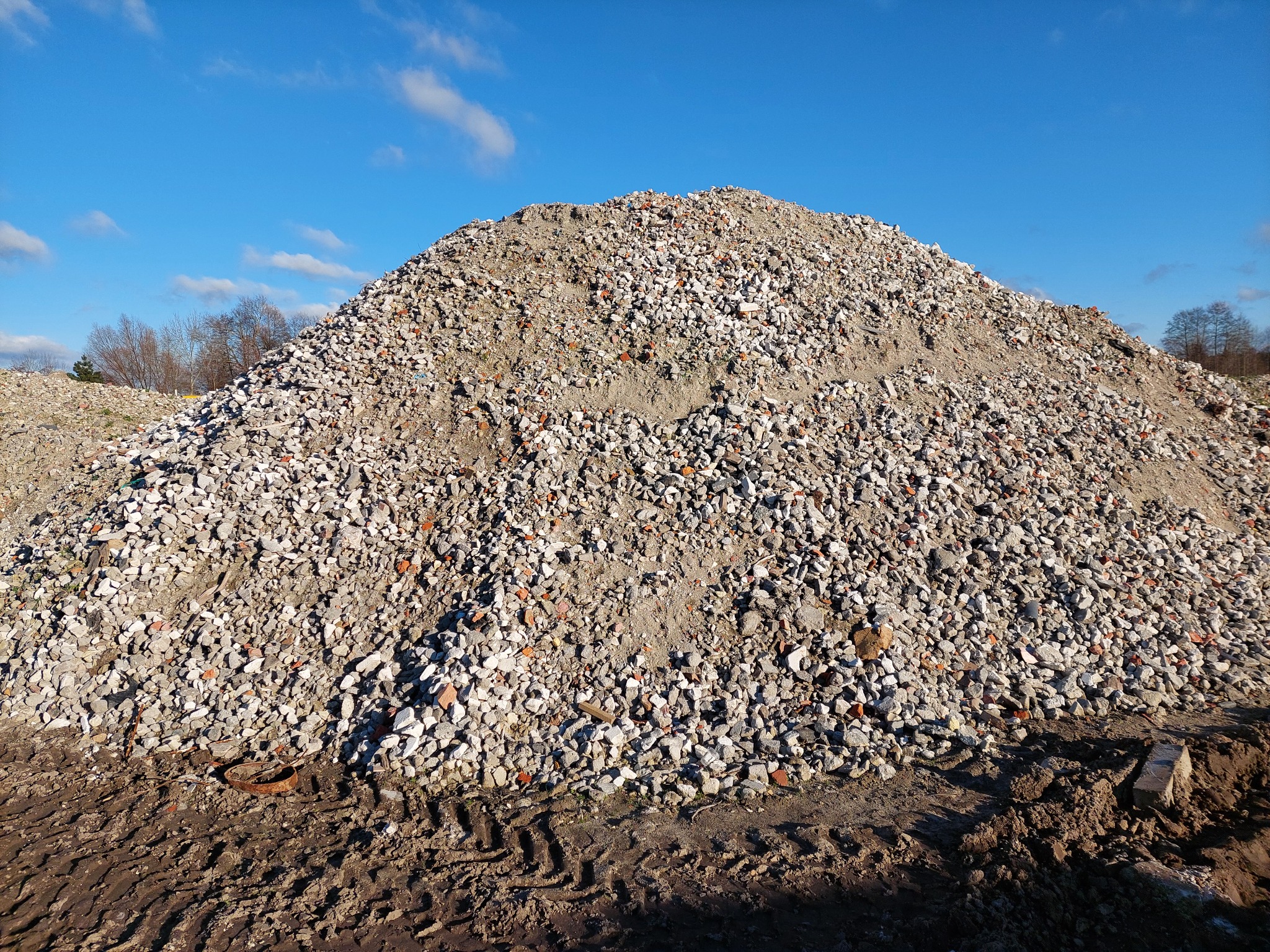 Góra gruzu ceglanego i kamiennego na ziemi z widocznymi śladami opon, błękitne niebo w tle.