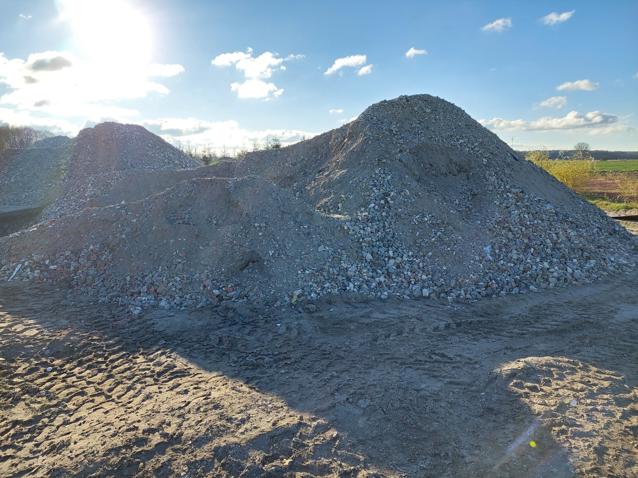Dwie pryzmy gruzu i ziemi na placu budowy, widoczne ślady opon pojazdów na ubitej ziemi, błękitne niebo z chmurami w tle.