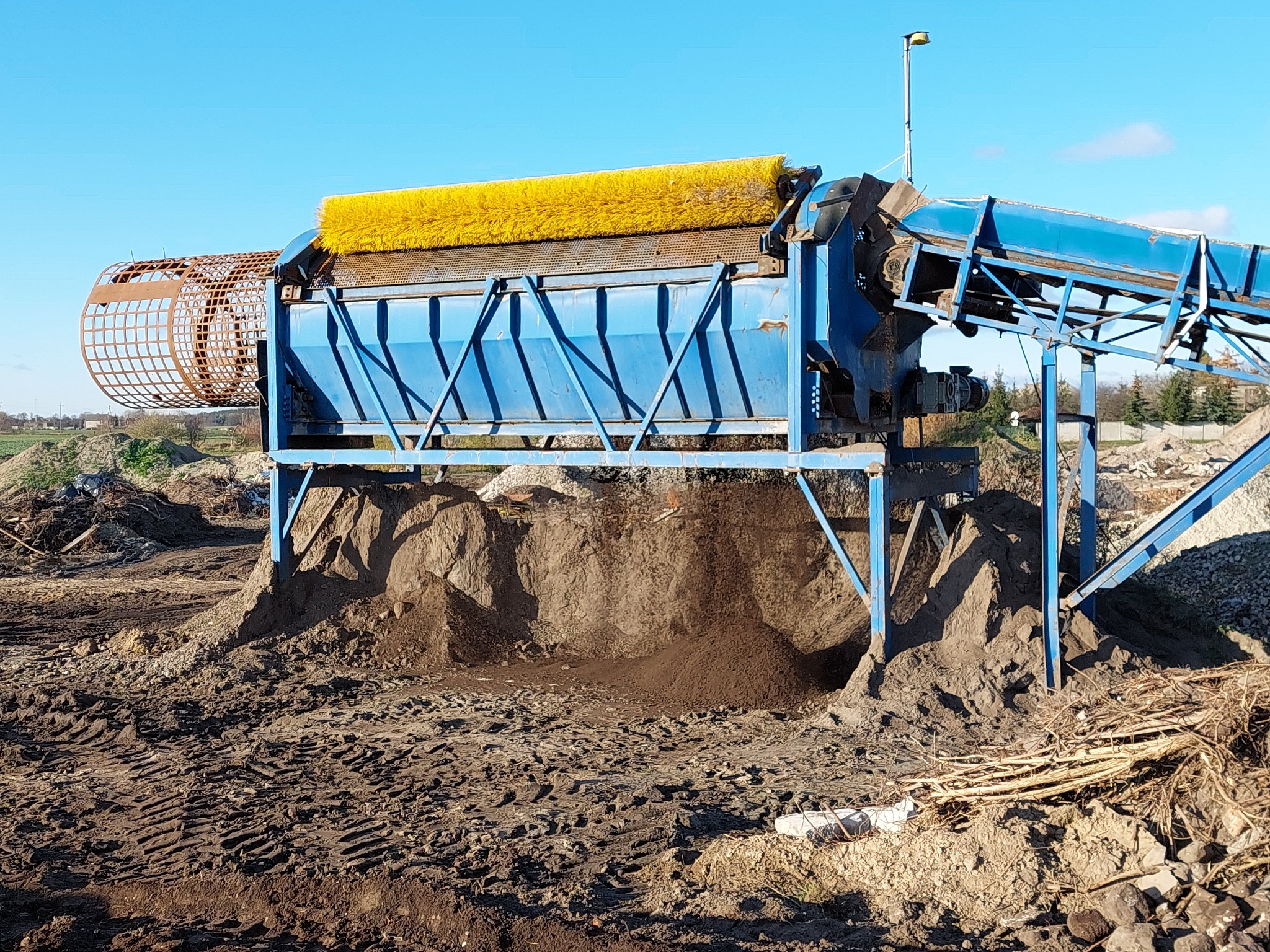 Niebieska przesiewaczka do ziemi z żółtą szczotką czyszczącą i cylindrycznym sitem, ustawiona na nierównym terenie z kopcami ziemi i śladami opon.
