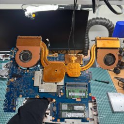 PIK Piotr Dziemiński - Płyta główna laptopa z systemem chłodzenia w dłoni serwisanta, widoczne elementy elektroniczne i radiator. Sprzęt na macie serwisowej, oświetlenie warsztatowe w tle.