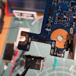 PIK Piotr Dziemiński - Płyta główna telefonu z odlutowanym gniazdem USB, trzymanym pęsetą. Widoczne ślady lutowania. Tło: mata serwisowa, narzędzia.