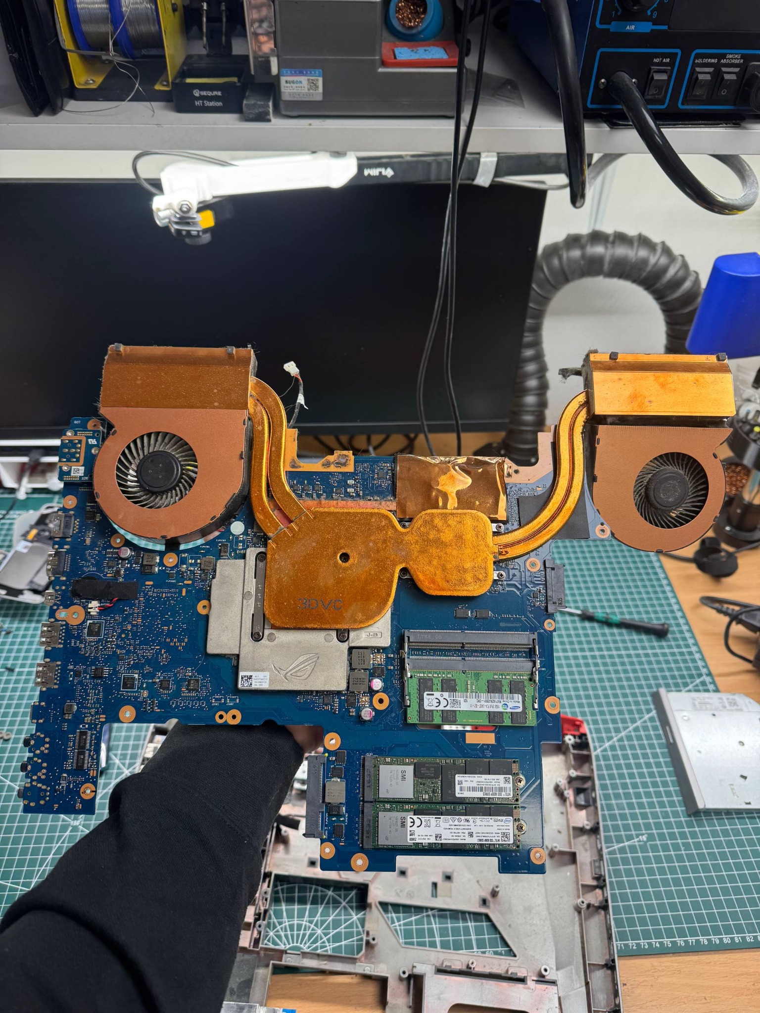 Płyta główna laptopa z systemem chłodzenia w dłoni serwisanta, widoczne elementy elektroniczne i radiator. Sprzęt na macie serwisowej, oświetlenie warsztatowe w tle.