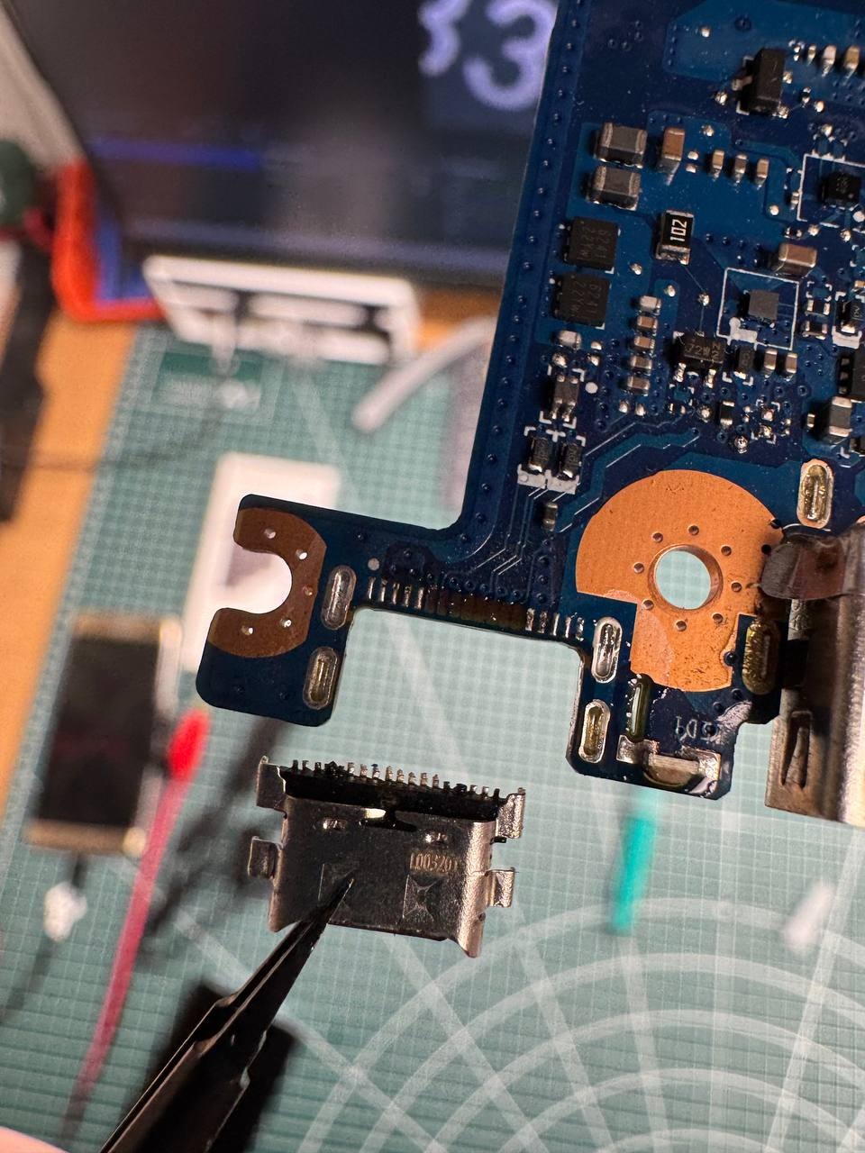Płyta główna telefonu z odlutowanym gniazdem USB, trzymanym pęsetą. Widoczne ślady lutowania. Tło: mata serwisowa, narzędzia.