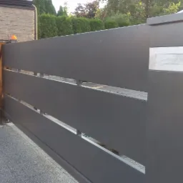 Nowoczesna brama wjazdowa w kolorze grafitowym z poziomymi panelami i skrzynką na listy, obok furtka do ogrodu w murze z cegły.