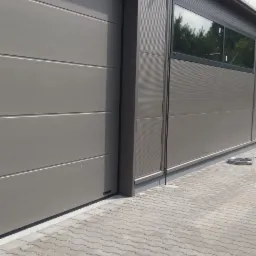 Szara segmentowa brama garażowa, część elewacji budynku z oknami, wyłożony kostką brukową podjazd.