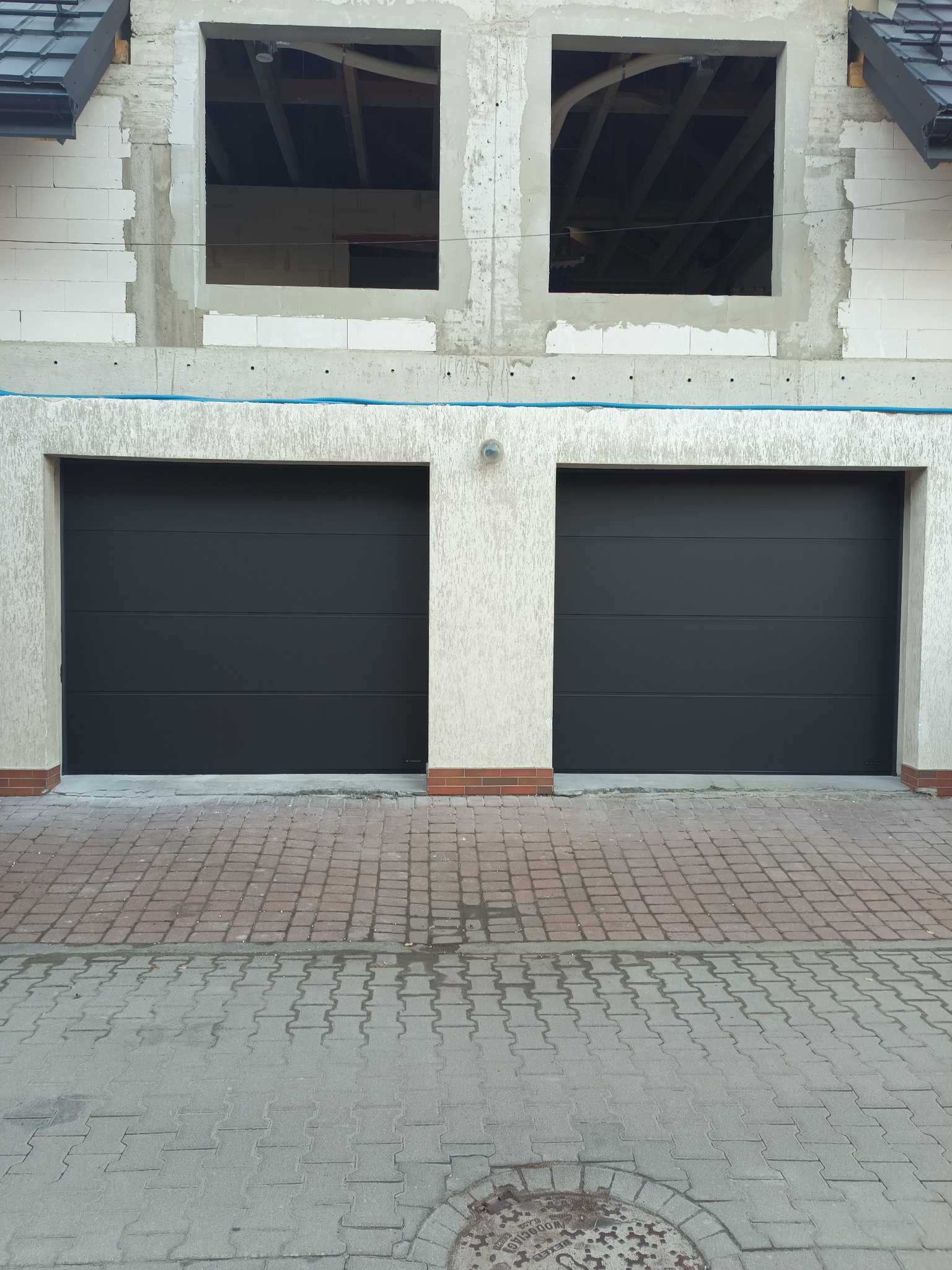 Dwie czarne bramy garażowe segmentowe w Podegrodziu, zamontowane w surowym budynku z cegły. Widoczne dwa otwory okienne na górze. Kostka brukowa przed budynkiem.