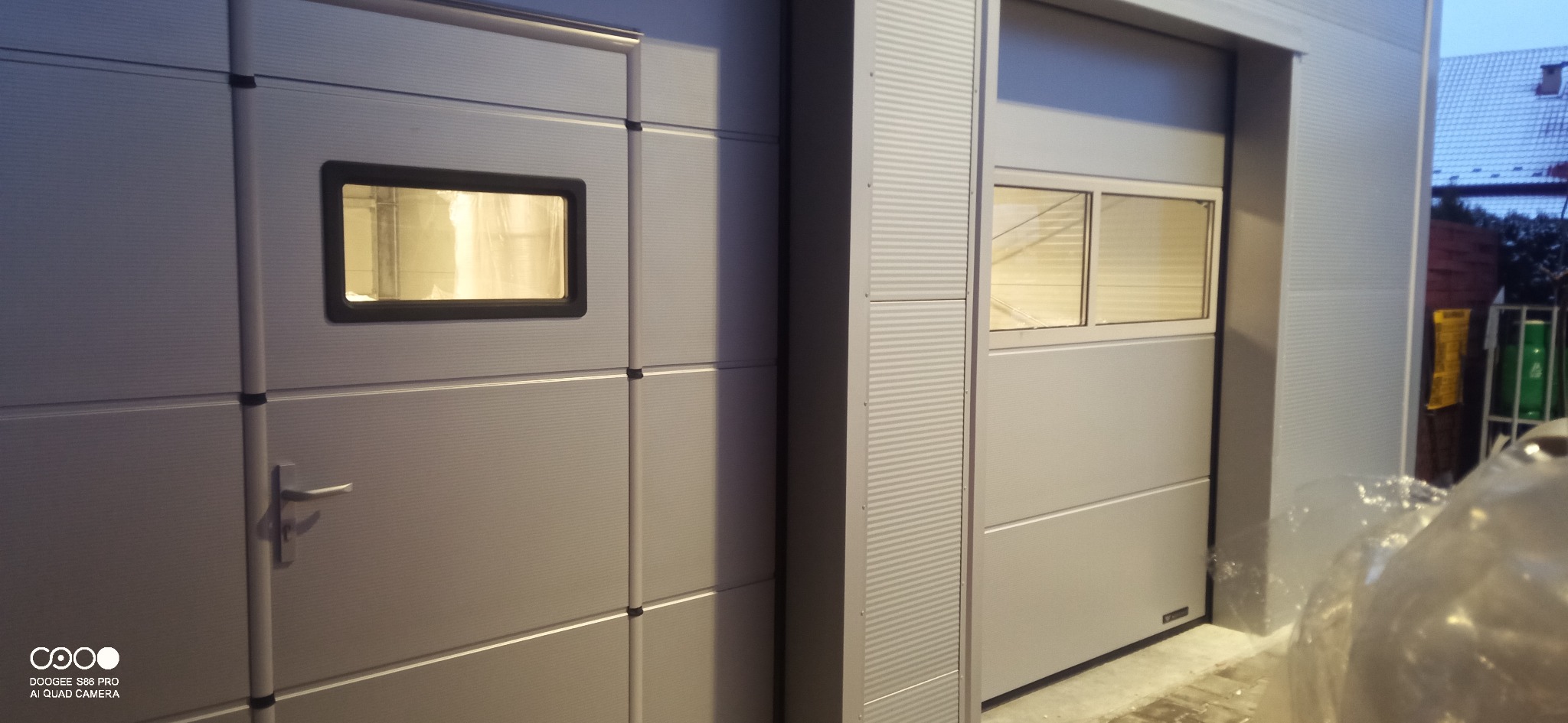 Dwie segmentowe bramy garażowe w kolorze srebrnym, z oknami. Jedna brama z uchwytem, druga z dwoma oknami. Widoczne elementy konstrukcyjne budynku.