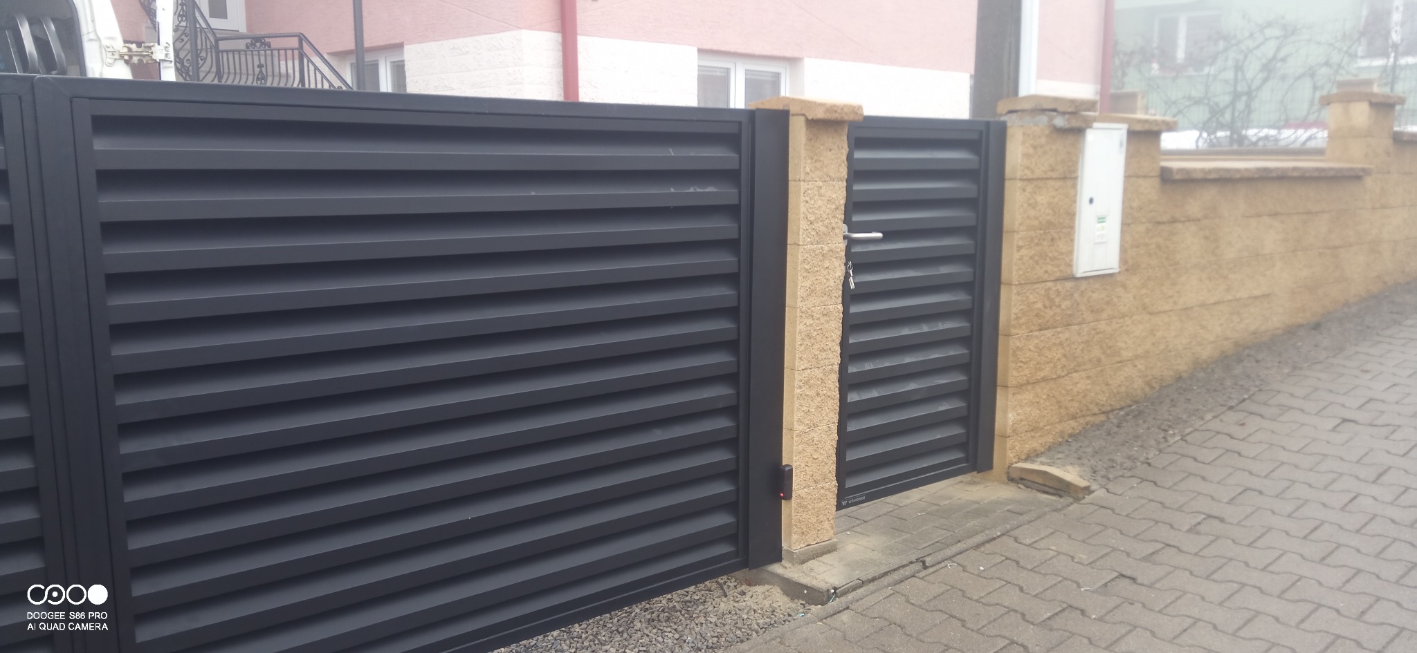 Nowoczesna, grafitowa brama wjazdowa lamelowa z furtką, osadzona na słupkach z piaskowca. Widok z zewnątrz na kostkę brukową i fragment domu w tle.