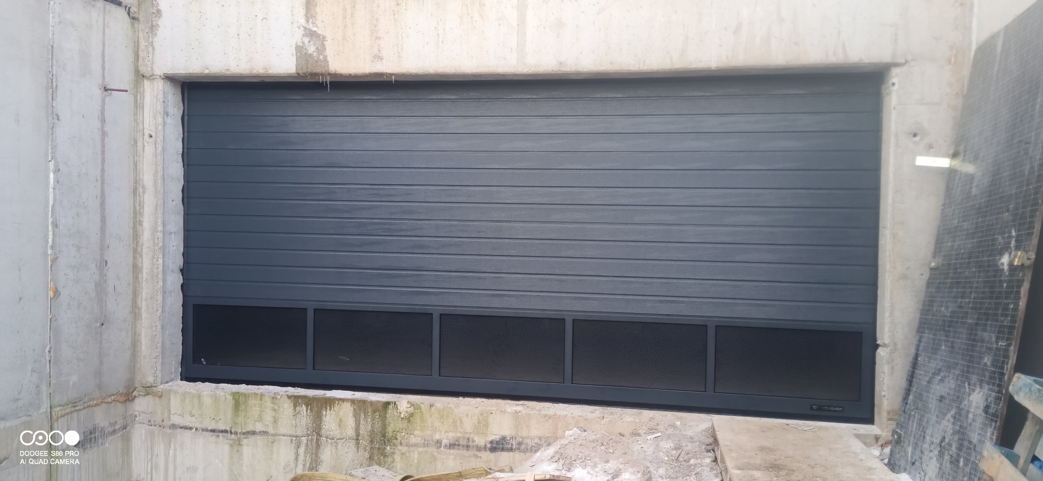 Szara brama garażowa segmentowa z kratką wentylacyjną w surowym otworze betonowym. Widoczne ślady montażu i zabrudzenia na betonie.