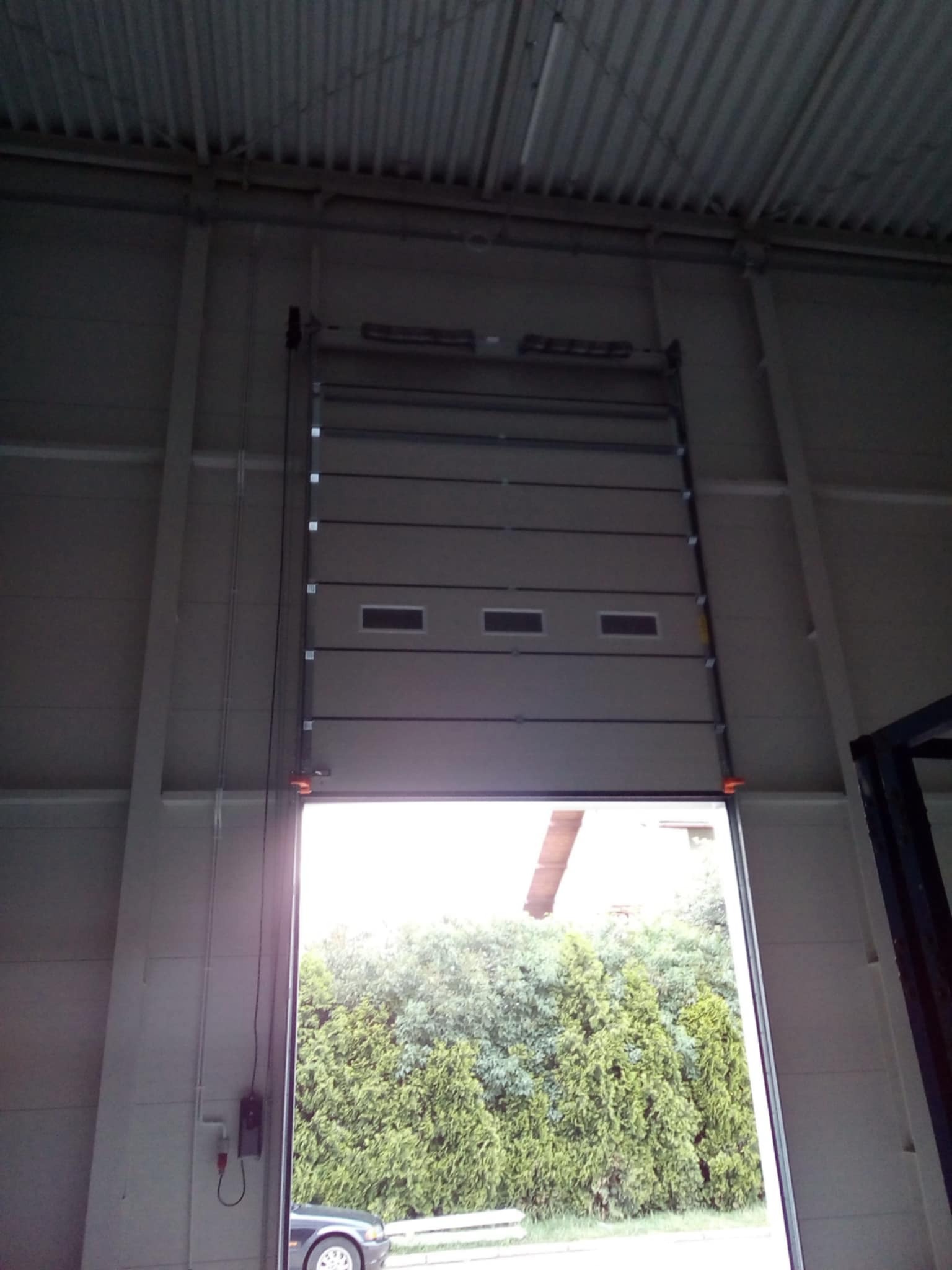 Otwarta segmentowa brama garażowa z oknami, widok z wnętrza hali na zieleń i fragment samochodu na zewnątrz. Widoczne prowadnice i mechanizm bramy.