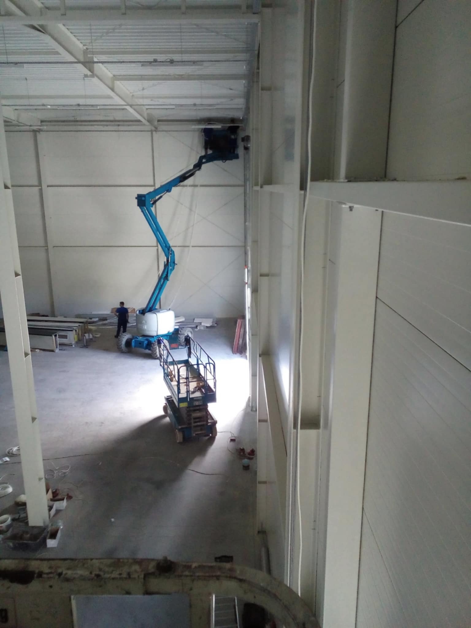 Wnętrze hali w trakcie montażu. Podnośnik koszowy z pracownikiem przy ścianie. Widoczne elementy konstrukcyjne i oświetlenie. Hala w trakcie budowy lub modernizacji.