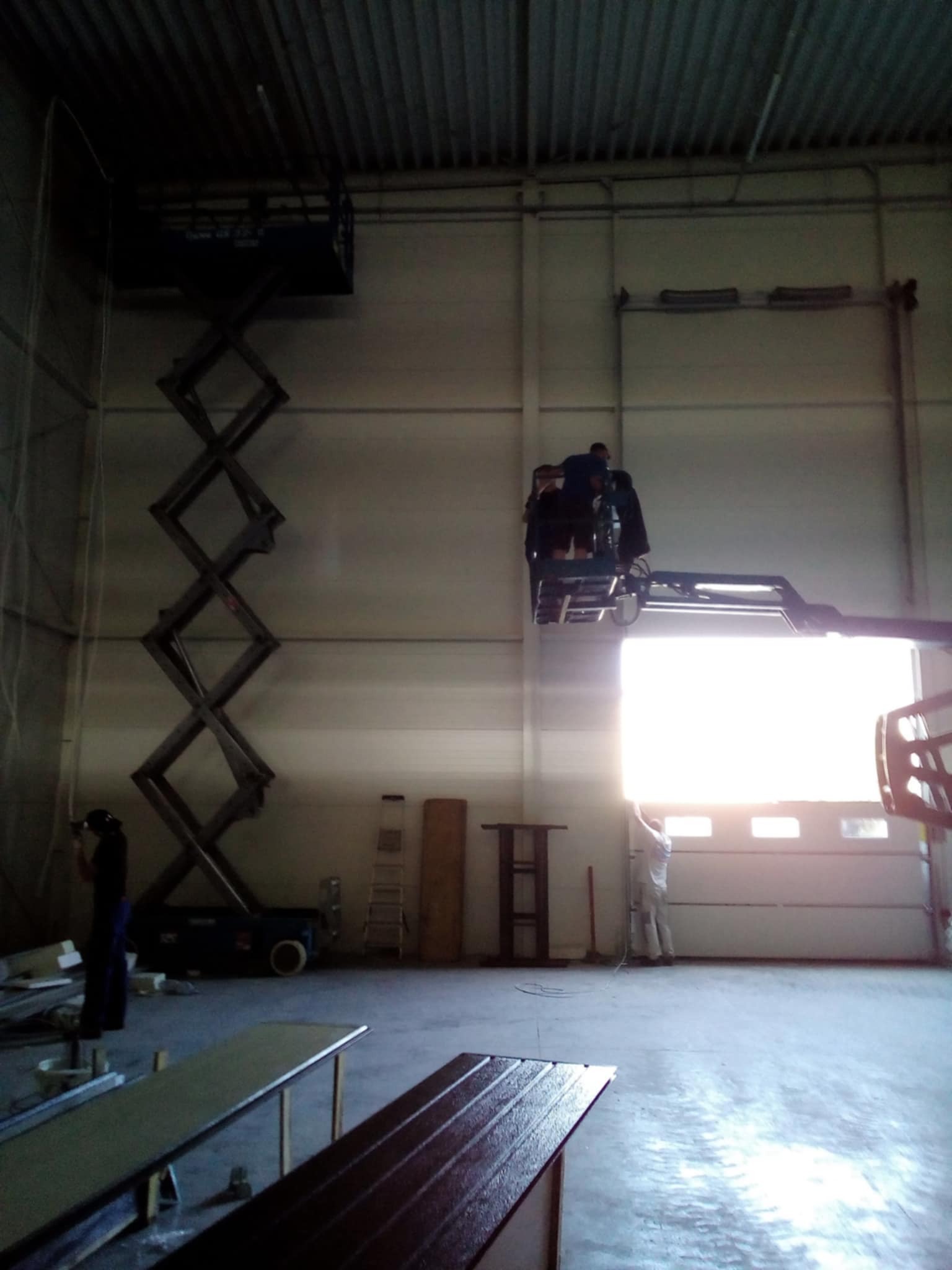 Montaż bramy garażowej segmentowej z podnośnika nożycowego w hali. Pracownicy montują bramę, a jeden z nich stoi na podłodze. Widoczne elementy bramy.