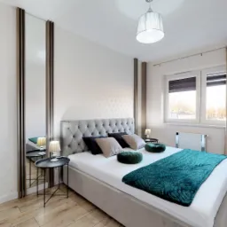 Elegancka sypialnia z łóżkiem kontynentalnym, szarą tapicerowaną ramą, turkusową narzutą, symetrycznie ustawionymi stolikami nocnymi i lustrami po bokach, jasnymi ścianami z subtelnym wzorem...