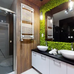 Nowoczesna łazienka z kabiną prysznicową, umywalkami nablatowymi, grzejnikiem drabinkowym i ścianą pokrytą mchem dekoracyjnym.