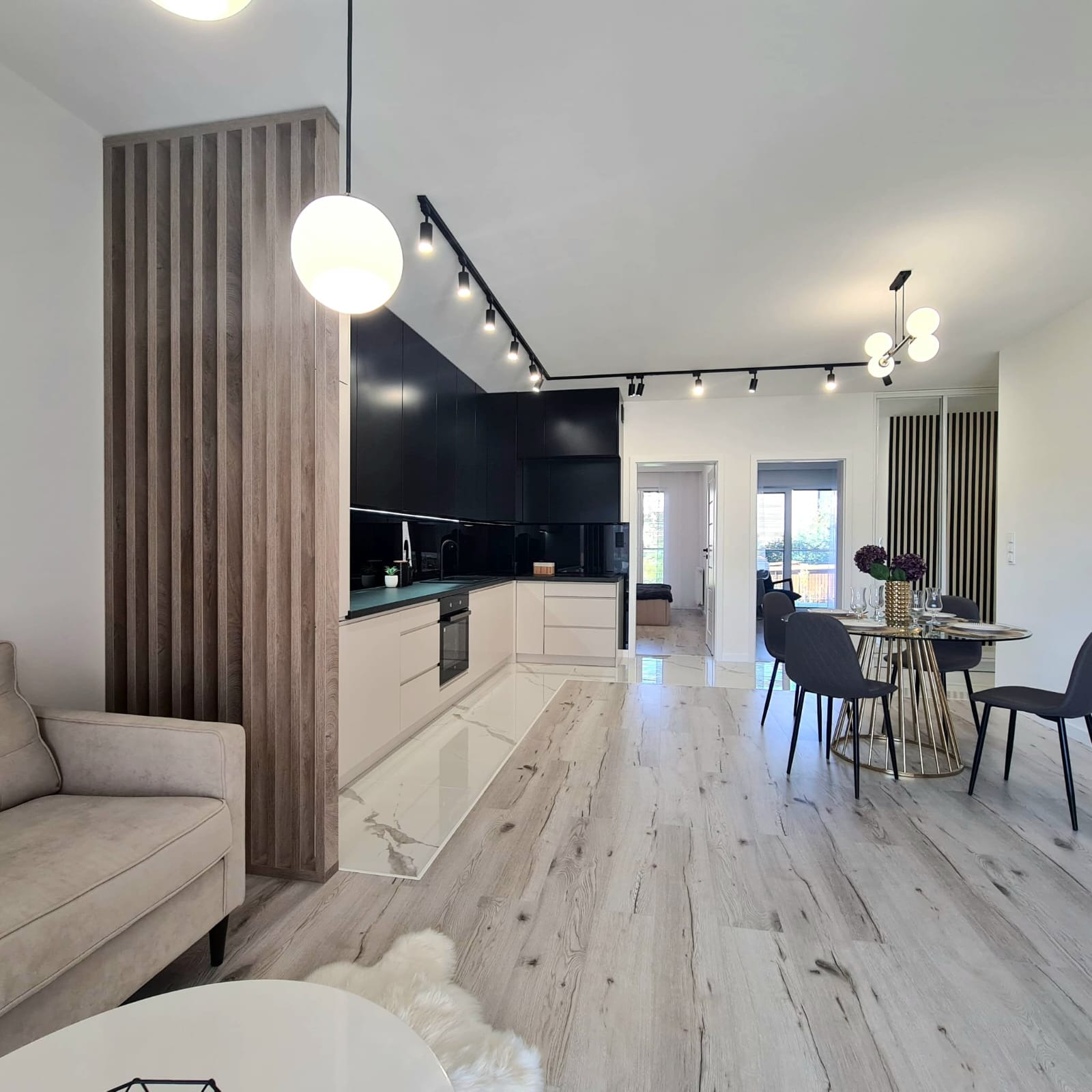 Nowoczesne wnętrze mieszkania z otwartą kuchnią, drewnianą ścianką działową, oświetleniem szynowym i okrągłym stołem jadalnym.