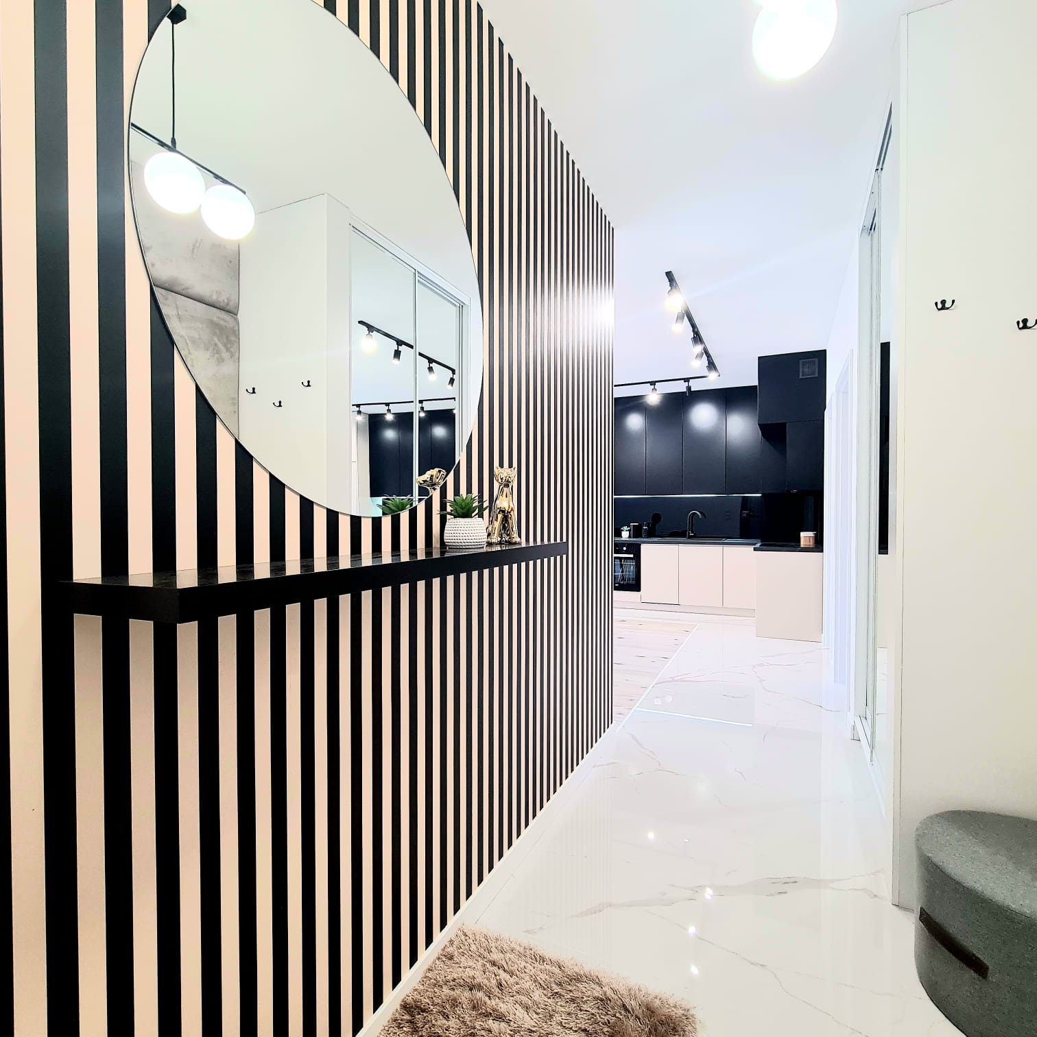 Nowoczesny przedpokój w Kielcach z tapetą w czarno-białe pasy, okrągłym lustrem, małą półką z dekoracjami i marmurową podłogą, prowadzący do jasnej kuchni.