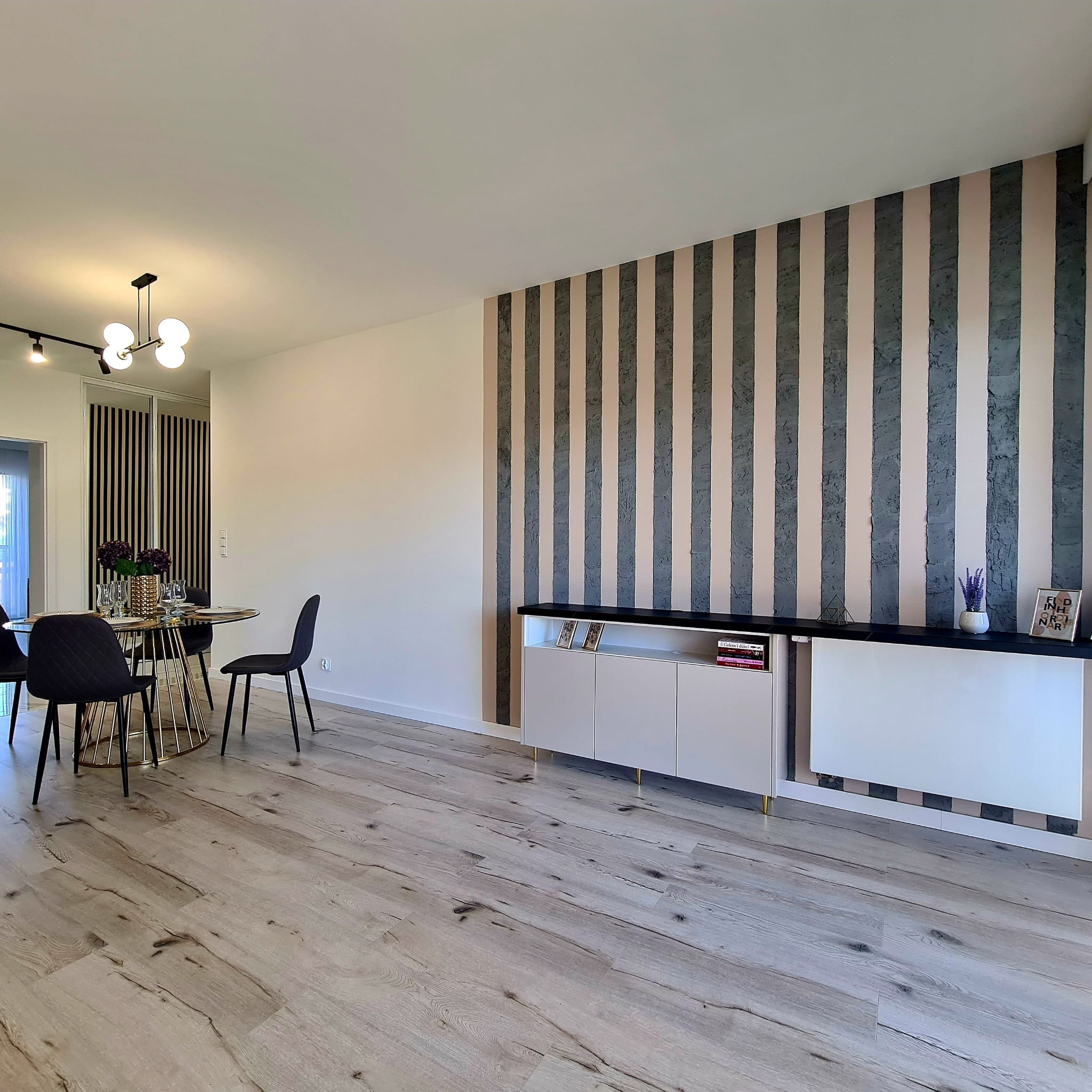 Jasne wnętrze jadalni z dekoracyjną ścianą w pionowe pasy, okrągłym stołem na złotej podstawie i drewnianą podłogą. Widoczny niski regał z dekoracjami na blacie.