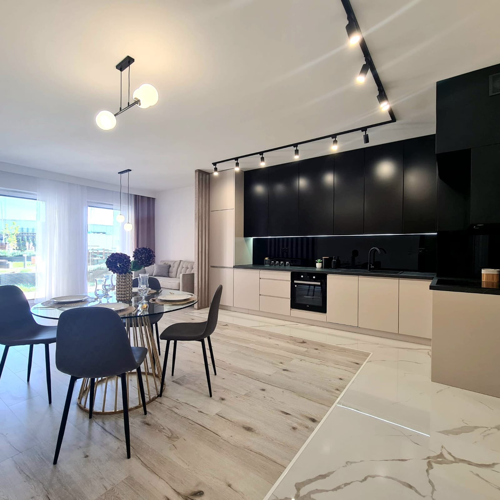 Nowoczesna kuchnia z czarnymi górnymi szafkami i beżowymi dolnymi, oświetlenie szynowe, okrągły stół z kwiatami i czarnymi krzesłami, widok na salon w tle.
