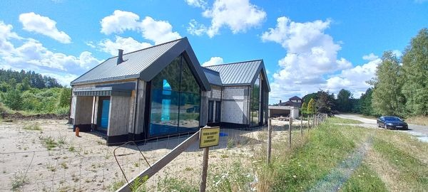 Nowoczesny dom jednorodzinny z dużymi przeszkleniami i dachem dwuspadowym, widoczny z zewnątrz, z żółtą tabliczką ostrzegawczą na pierwszym planie i samochodem na drodze w tle.