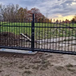 Czarna, metalowa brama przesuwna w trakcie montażu na betonowej podstawie, widoczny nasyp ziemi i elementy konstrukcyjne, tło stanowią trawnik i drzewa w jesiennej scenerii.