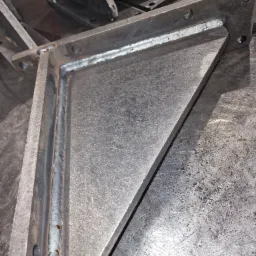 Szara, metalowa konstrukcja spawana w kształcie kątownika z otworami montażowymi, leżąca na metalowym stole warsztatowym, widoczne ślady spawania i obróbki metalu.