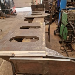 Uslugi Spawalniczo-Monterskie - Surowe, metalowe elementy konstrukcyjne z owalnymi otworami, spawane na metalowych kozłach w warsztacie, w tle wózek widłowy i butla z gazem spawalniczym.