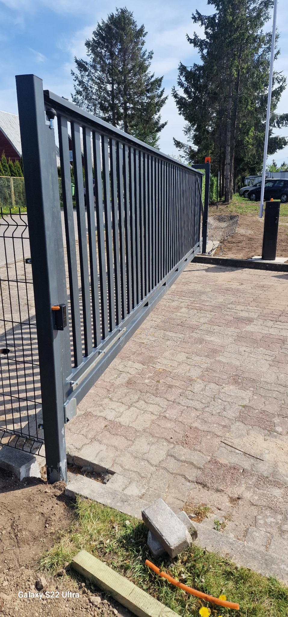 Nowoczesna, przesuwna brama wjazdowa z automatyką, widoczna szyna zębatkowa, na posesji wyłożonej kostką, z elementami trawnika i ogrodzenia siatkowego.