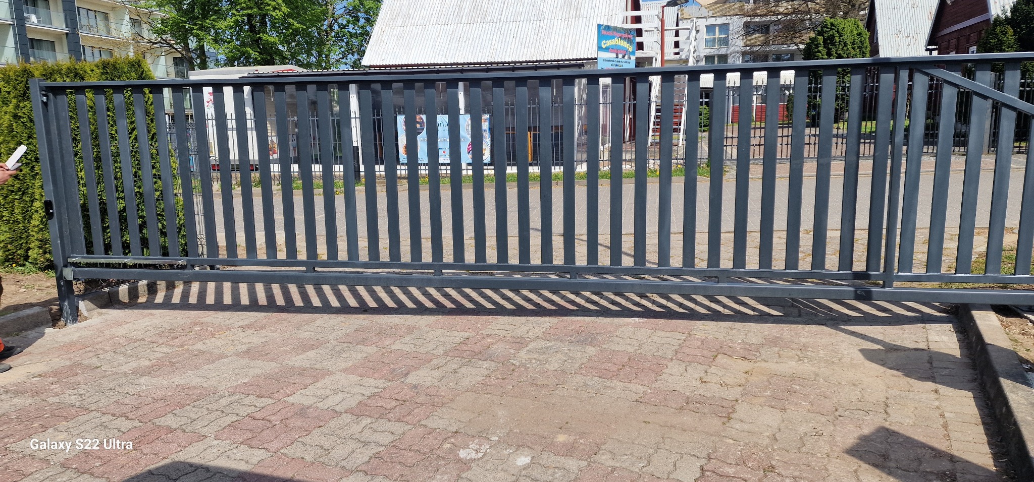 Szara, przesuwna brama wjazdowa z pionowymi szczeblami, widok z zewnątrz na kostkę brukową i budynki w tle.