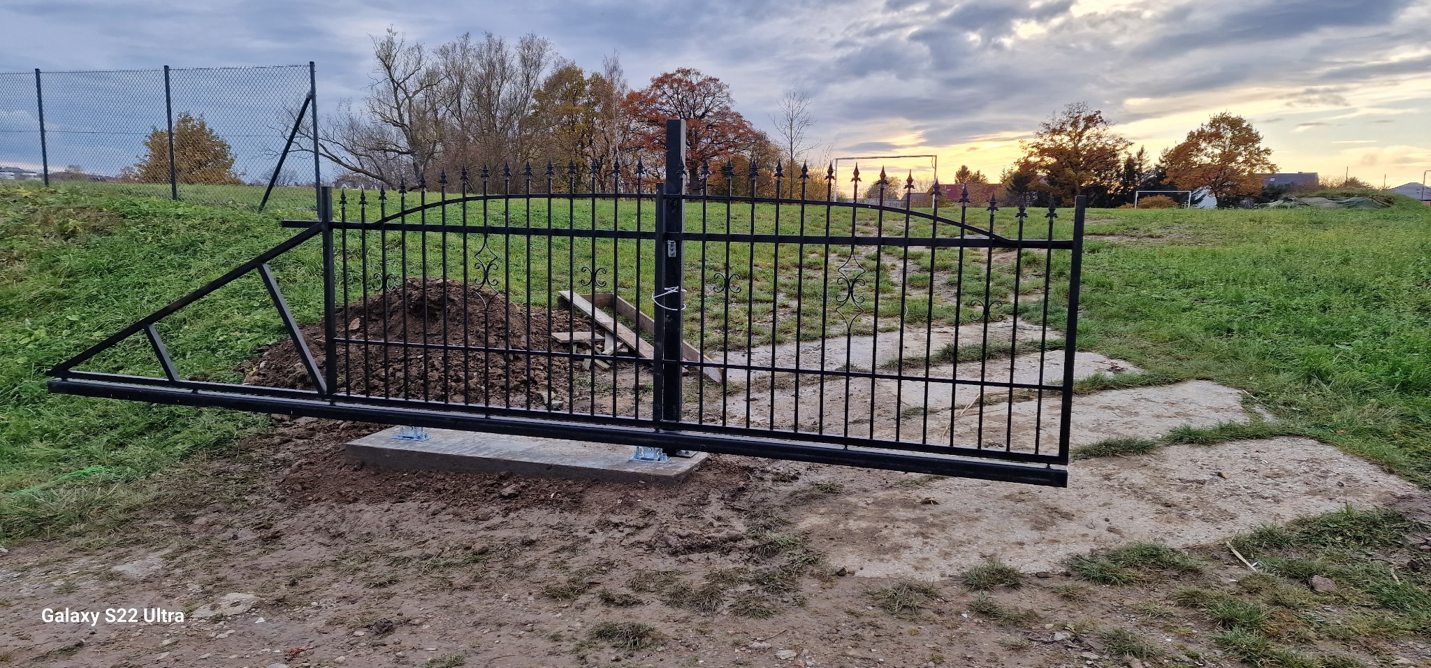 Czarna, metalowa brama przesuwna w trakcie montażu na betonowej podstawie, widoczny nasyp ziemi i elementy konstrukcyjne, tło stanowią trawnik i drzewa w jesiennej scenerii.