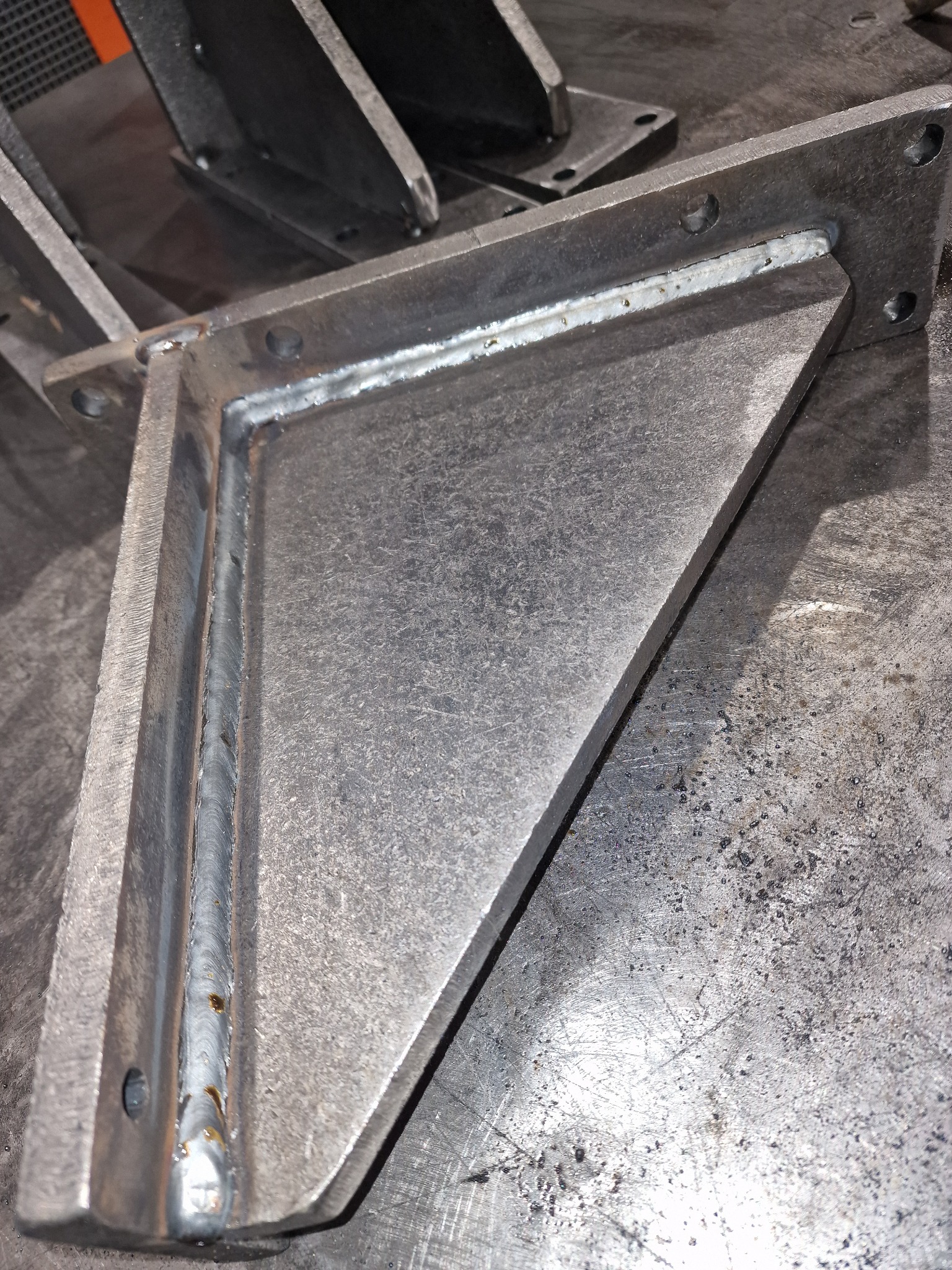 Szara, metalowa konstrukcja spawana w kształcie kątownika z otworami montażowymi, leżąca na metalowym stole warsztatowym, widoczne ślady spawania i obróbki metalu.