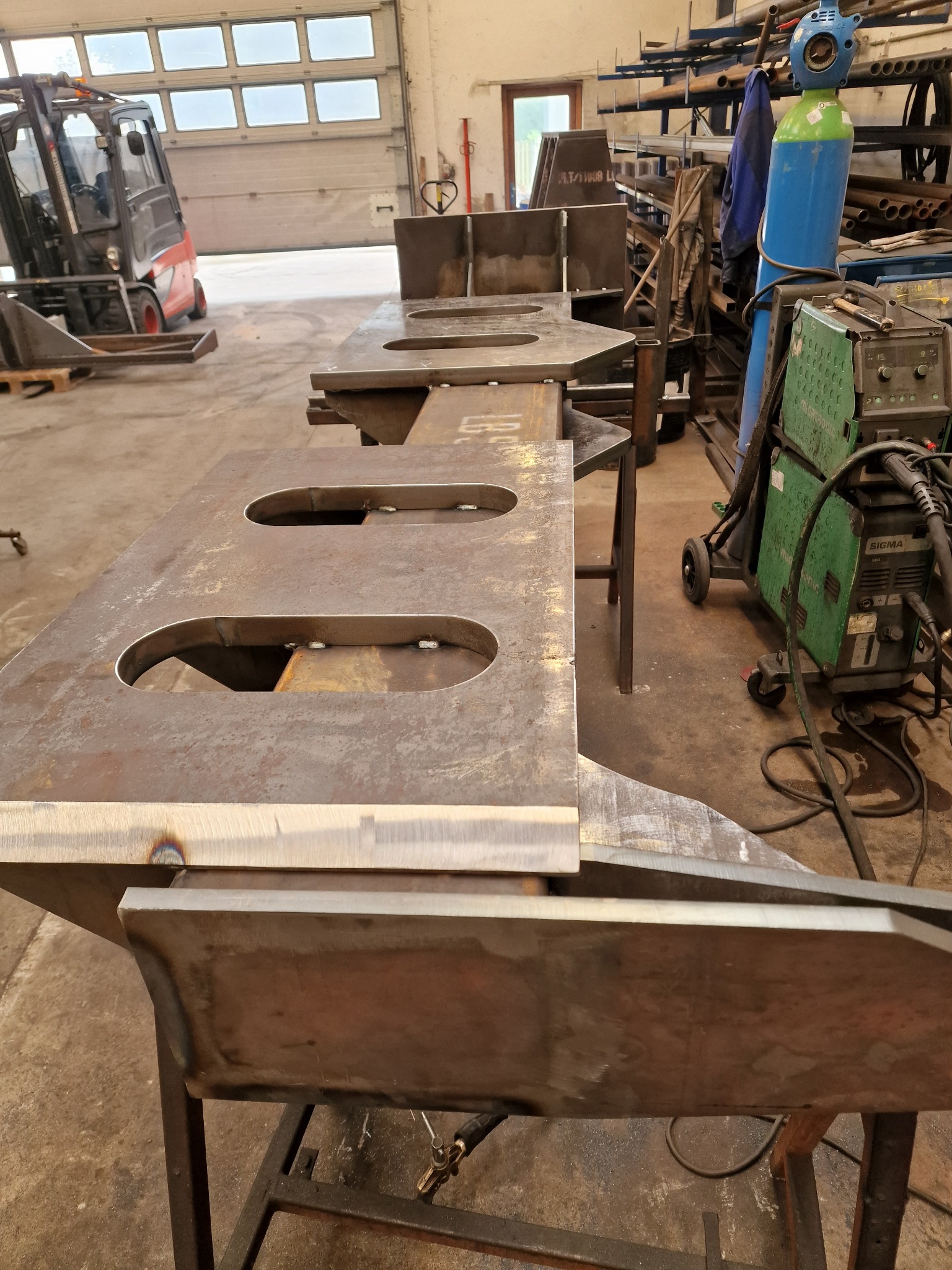 Surowe, metalowe elementy konstrukcyjne z owalnymi otworami, spawane na metalowych kozłach w warsztacie, w tle wózek widłowy i butla z gazem spawalniczym.