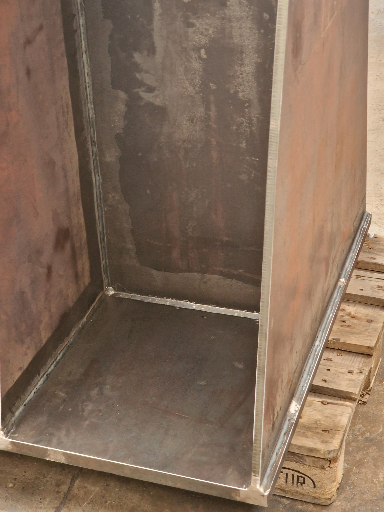 Surowa, spawana metalowa konstrukcja w kształcie prostopadłościanu bez jednej ściany, umieszczona na drewnianej palecie, widoczne spawy.