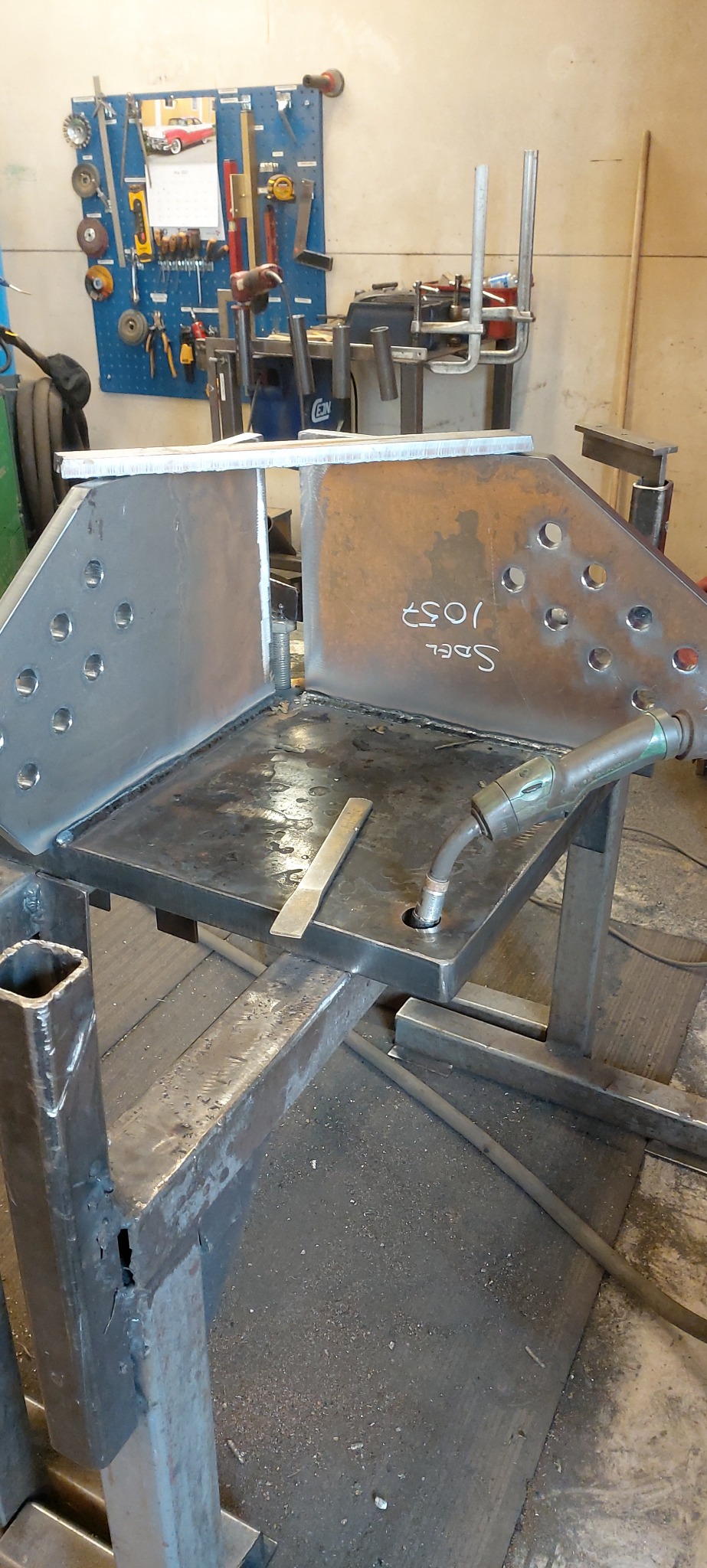 Konstrukcja metalowa w trakcie spawania, widoczny uchwyt spawalniczy, metalowe profile i blachy z otworami. Na jednej z blach napis 'Sdel 1057'. W tle tablica z narzędziami.