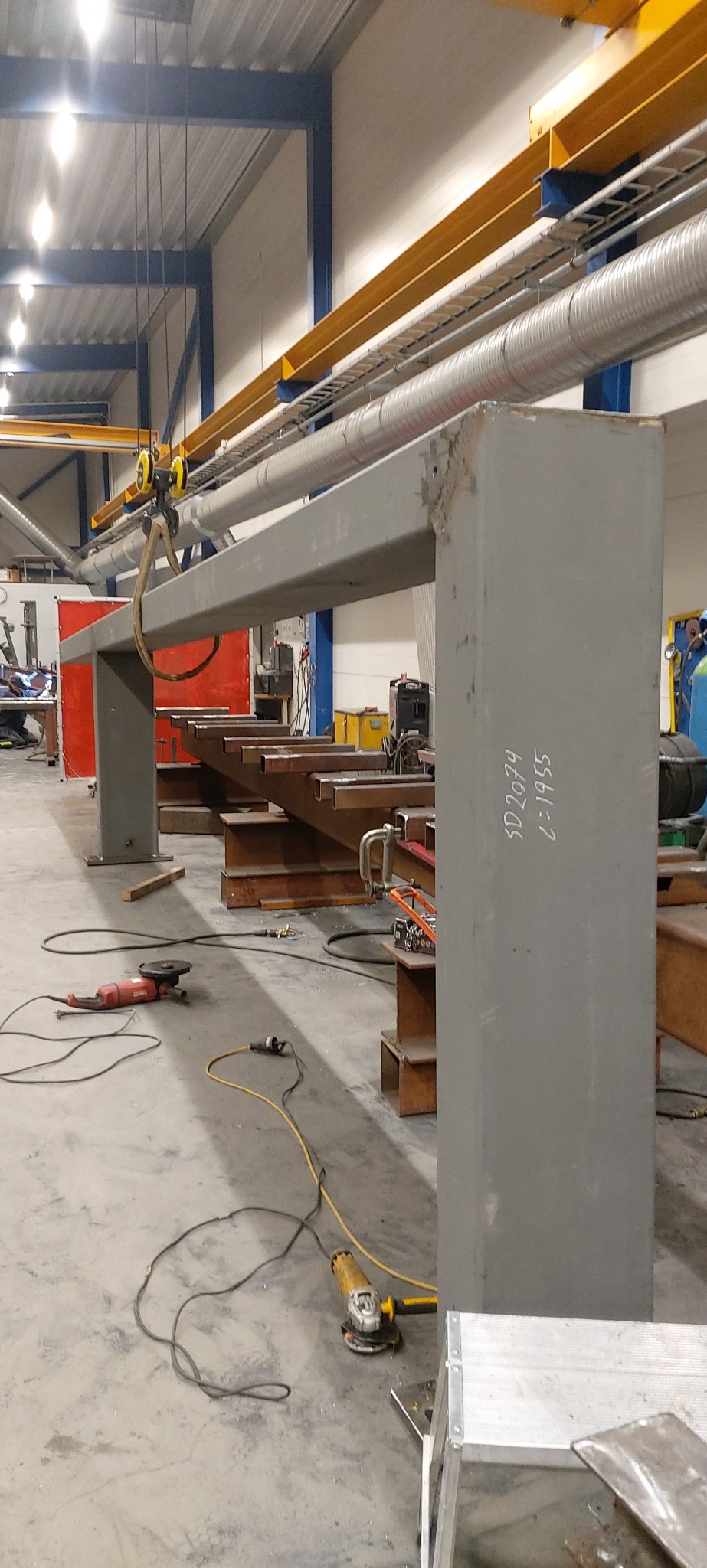 Szara, spawana konstrukcja metalowa w hali produkcyjnej, z widocznym suwnicą, narzędziami szlifierskimi, spawarką i fragmentami schodów metalowych w tle; na elemencie konstrukcji widoczne napisy SD...