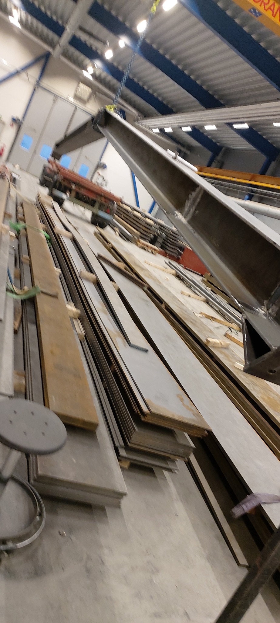 Stalowe płyty i profile przygotowane do obróbki, widoczne elementy konstrukcyjne suwnicy w hali produkcyjnej.