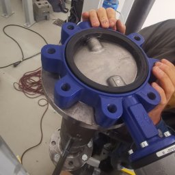 HR-MONT Service - Monteraż zaworu motylkowego, niebieska obudowa, metalowa tarcza, widoczne śruby mocujące, instalacja w pomieszczeniu technicznym.