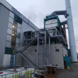 Zewnętrzny widok przemysłowej instalacji z metalową konstrukcją schodów i wysokim kominem, zbiorniki z płynem na pierwszym planie, pochmurne niebo w tle.