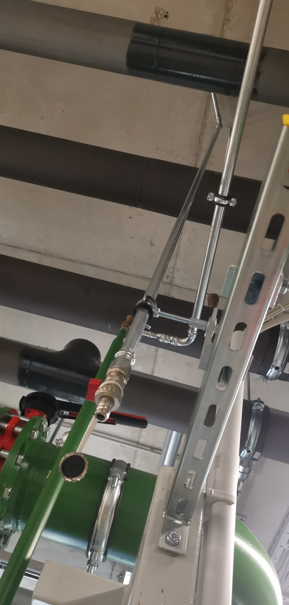 Fragment instalacji hydraulicznej z rurami, zaworami i elementami mocującymi, widoczny na tle sufitu z rurami.