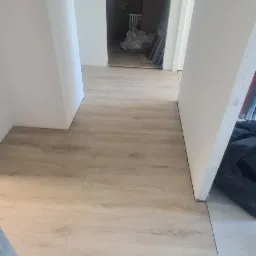 Korytarz po remoncie z nową, jasną podłogą imitującą drewno, widoczne drzwi do innego pomieszczenia w trakcie prac wykończeniowych.