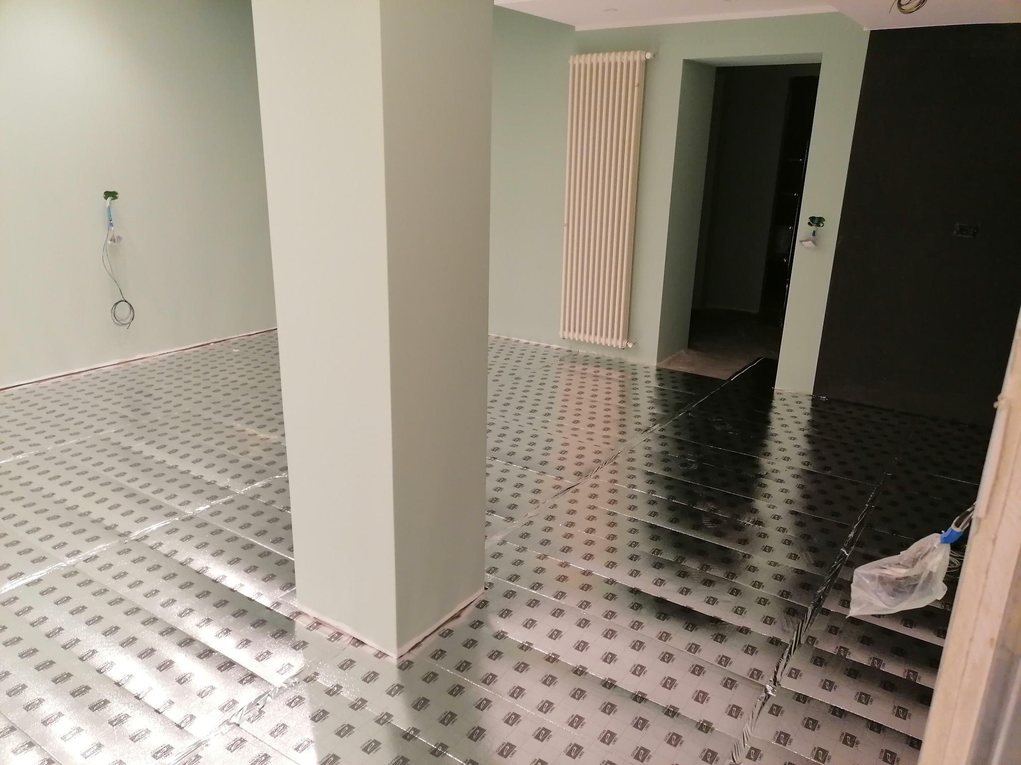 Rozłożona folia grzewcza na podłodze w pomieszczeniu z jasnymi ścianami, widoczny grzejnik i przygotowane przyłącze elektryczne.