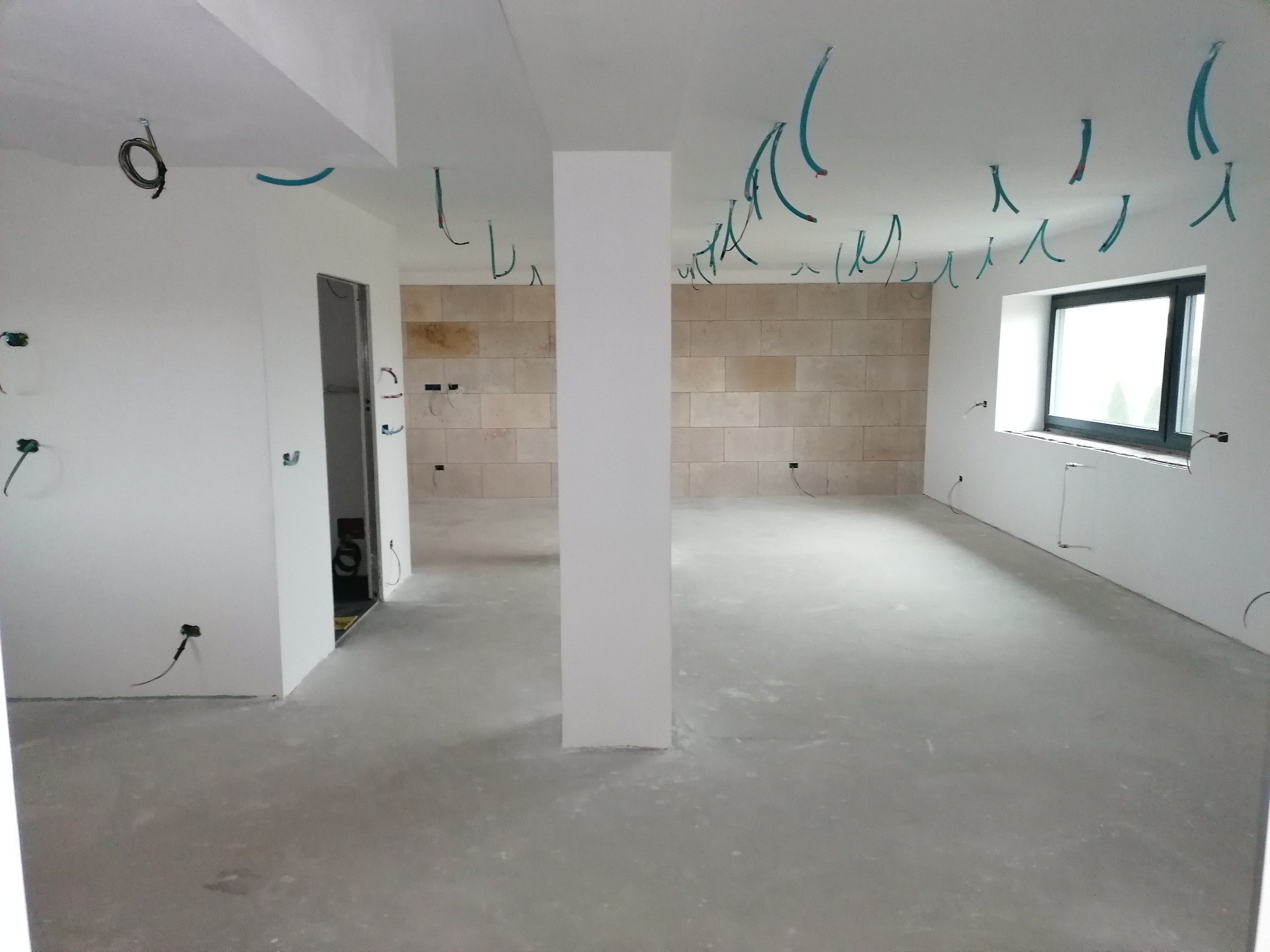 Wnętrze w trakcie prac wykończeniowych: surowy beton na podłodze, ściany częściowo wykończone płytkami imitującymi kamień, wystające przewody elektryczne z sufitu i ścian, widoczne puszki...