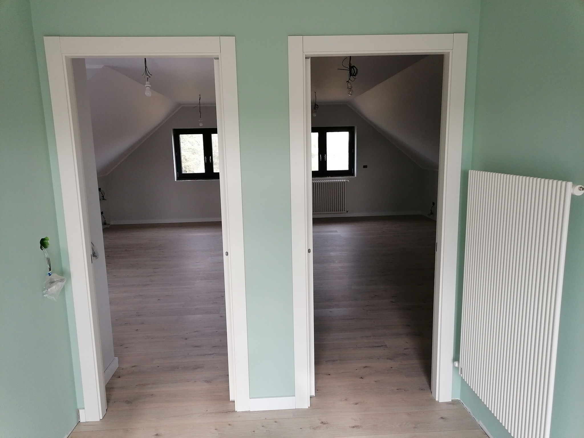 Dwa białe, minimalistyczne ościeżnice drzwiowe w Lipno, prowadzące do poddaszowego wnętrza z drewnianą podłogą i grzejnikiem, ściany w kolorze miętowym, widoczne niedokończone instalacje elektryczne.