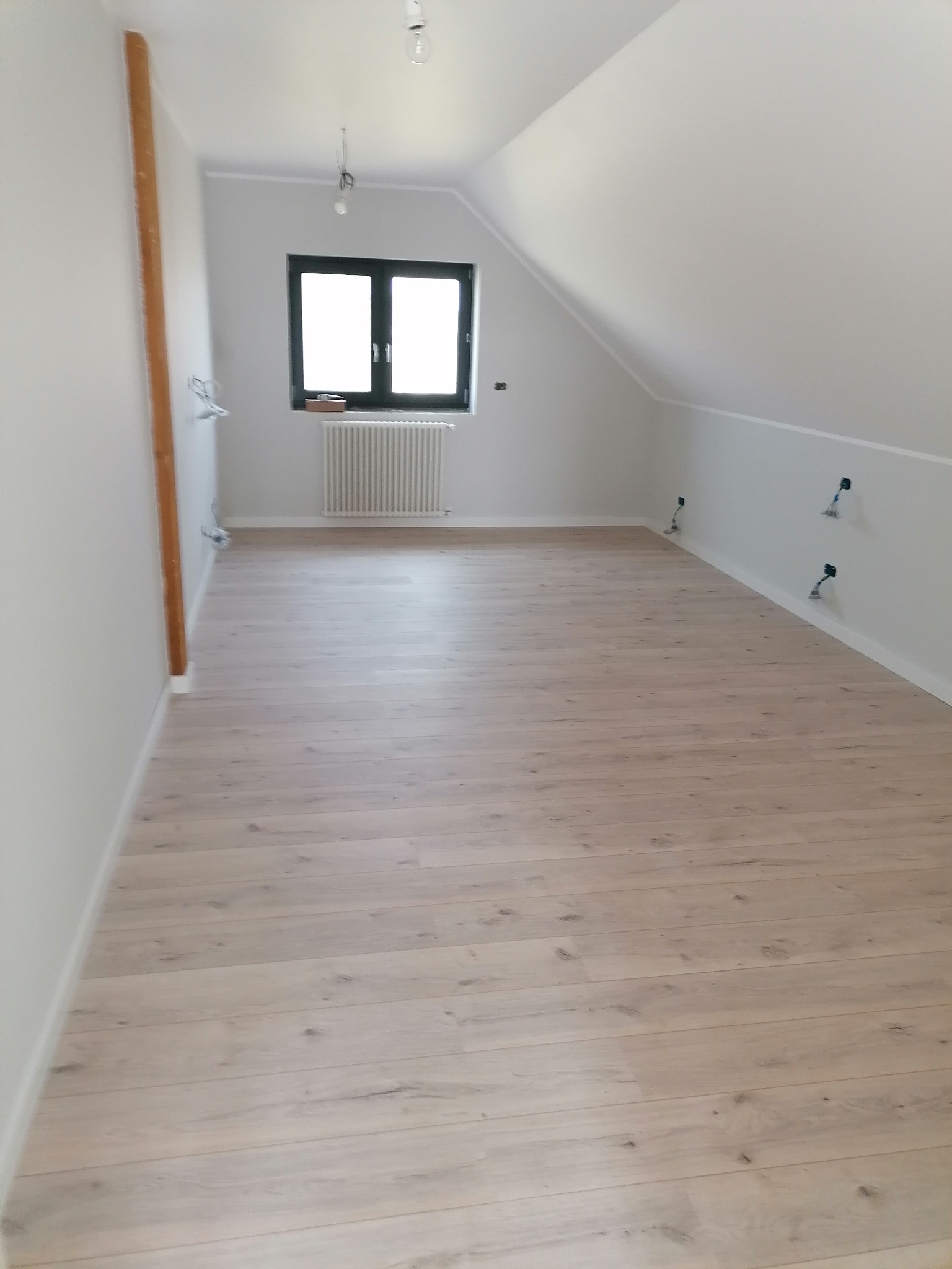 Poddasze z nową, jasną podłogą panelową, świeżo pomalowane ściany na biało, okno dachowe z czarną ramą i grzejnikiem pod nim, widoczne niedokończone instalacje elektryczne.