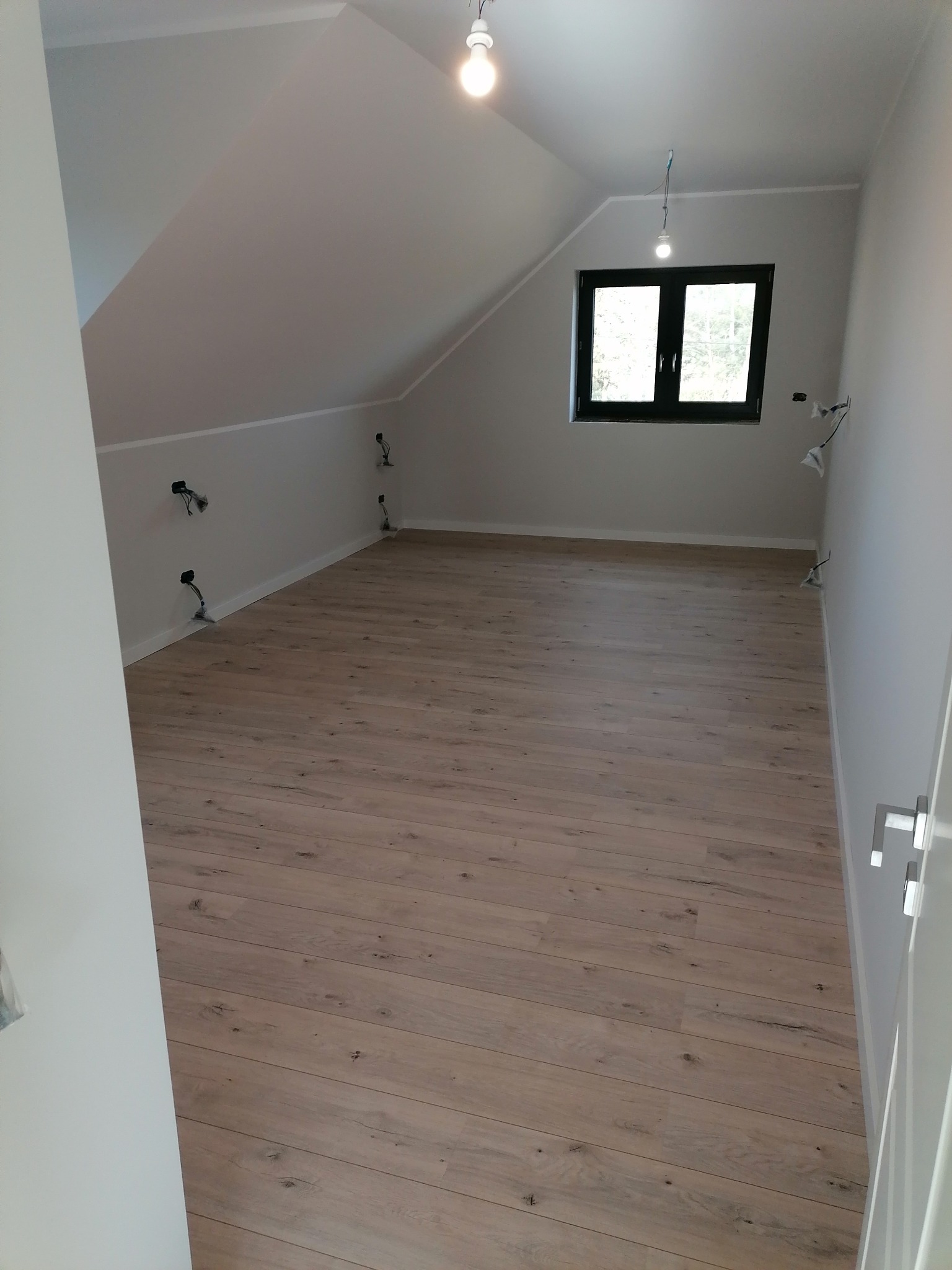 Poddasze z drewnianą podłogą w trakcie wykańczania, widoczne niedokończone punkty elektryczne na ścianie i surowa żarówka, czarne okno dachowe.